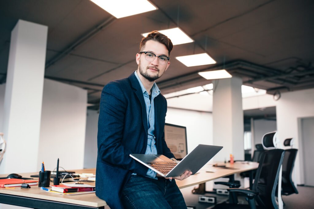 attractive man glassess is sitting near workplace office he wears blue shirt dark jacket he holds laptop looks camera
